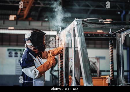 Industrial worker welds metal framework with sparks flying. Worker wears protective gear, focused expression. Factory setting shows skilled labor in m Stock Photo