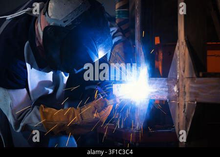 Welder in protective gear works closely on metal. Bright sparks fly from the welding torch. The worker's posture shows deep concentration. Industrial Stock Photo