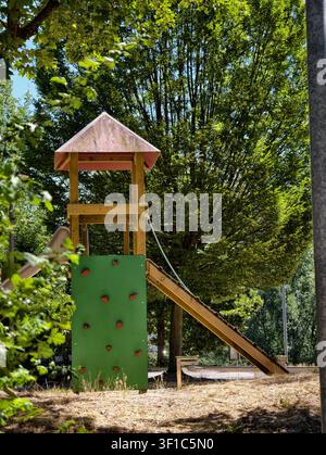 Germany 2026 Wooden playground climbing frame with slide tower stands in lush green park, surrounded by leafy trees, grass and bright summer sunlight, Stock Photo