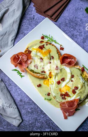 Eggs Benedict with hollandaise sauce, prosciutto and fresh herbs served on a white plate, overhead view. Stock Photo