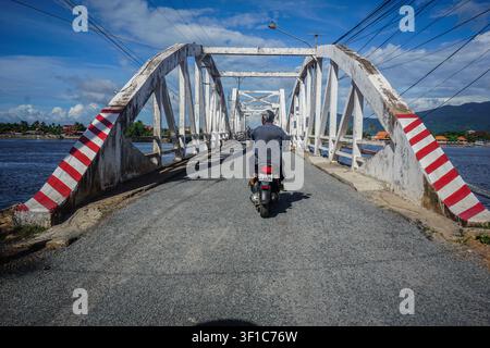 Kampot, Cambodia - September 02, 2016: A person wearing a gray shirt and dark pants rides a silver scooter away from the camera on a gray asphalt brid Stock Photo
