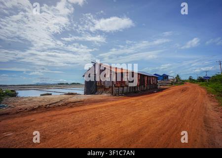 A weathered brown and rust-colored corrugated metal building stands on wooden stilts beside a wide, reddish-brown dirt road that curves into the dista Stock Photo