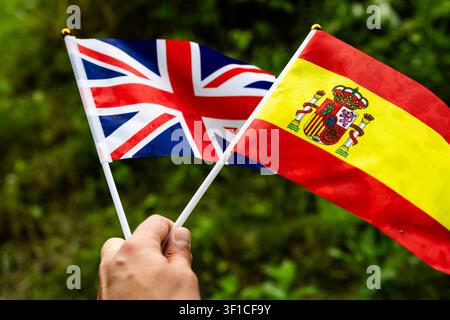 Hand holding two small flags against a green background showing the Union Jack of the United Kingdom and the yellow and red flag of Spain symbolizing Stock Photo