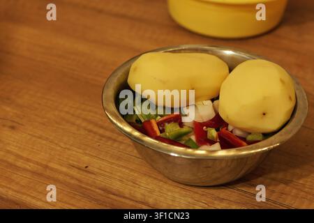 Peeled potatoes resting on a bed of freshly chopped red and green bell peppers and white onions in a stainless steel bowl, preparing for a homemade he Stock Photo