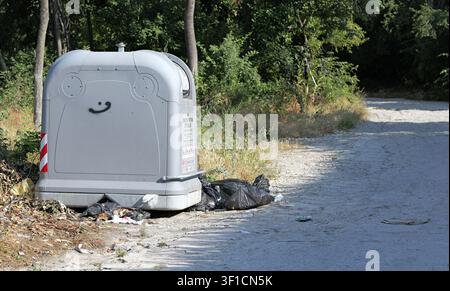 Grey plastic outdoor recycling bin standing beside a dirt road with green foliage. Showing a smiling face graffiti and black garbage bags on the groun Stock Photo