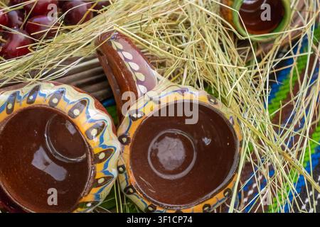 Traditional ceramic cups arranged with hay, cherries and colorful textile, creating a rustic Balkan market still life with handmade pottery detail. Stock Photo