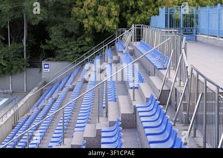 Long rows of empty blue plastic seats ascending upwards along concrete steps in an outdoor stadium or arena, featuring numerous metallic railings and Stock Photo
