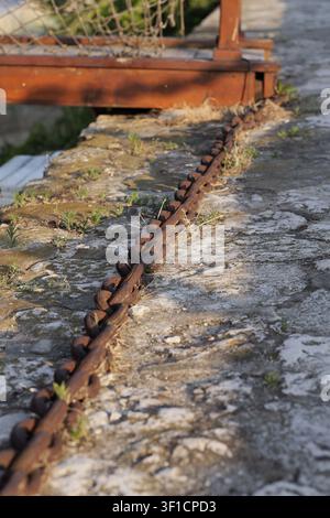Heavy rusted metal chain links are resting on an old textured stone path, showing signs of weathering and neglect with small plants growing through th Stock Photo