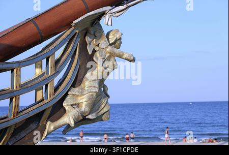 Golden figurehead sculpture on a wooden ship's bow standing on a sandy beach, with people enjoying the calm sea and blue sky in the background Stock Photo