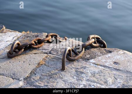 Rusty industrial chain resting on an old stone pier, providing mooring for boats in a harbor, connecting strength and security with the water backgrou Stock Photo