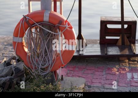 Orange life ring hanging with ropes on a weathered pole, symbolizing safety, rescue, and security by the water, offering protection at the port Stock Photo
