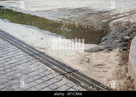 A large puddle of murky water reflects green trees on a cracked concrete surface adjacent to a metal grate and cobblestone path Stock Photo