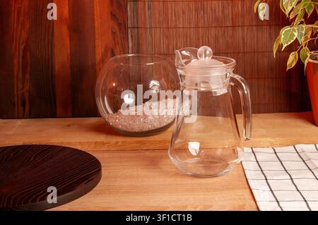 A transparent pitcher and a round glass bowl filled with small crystals are arranged on a rustic wooden table with a plant in the background Stock Photo