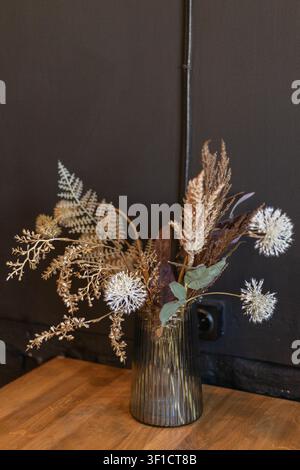 Detailed close up shot of an arrangement of dried flowers, fern leaves, and decorative plants inside a ribbed glass vase standing on a wooden table su Stock Photo