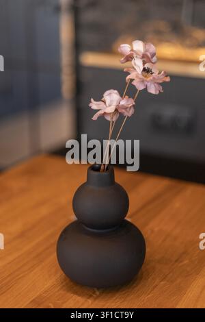 Detailed high angle shot of an arrangement of delicate pink faux flowers inside a matte black ceramic gourd vase standing on a wooden surface with a b Stock Photo