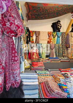 Paris, France - December 10, 2025: vibrant market stall is filled with colorful, patterned scarves, shawls, and textiles. Stacks of folded fabrics of Stock Photo
