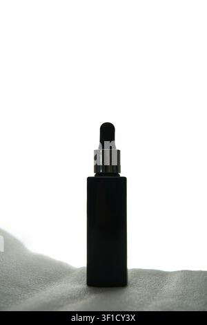 Matte Black Dropper Bottle Standing Upright On Soft Fabric Surface With Bright Backlight And Minimalist White Background For Cosmetic Packaging Display. Stock Photo