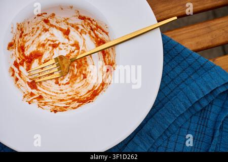 Empty plate with tomato sauce remains and fork on table beside cloth napkin. Concept of finished meal, food consumption and clean up Stock Photo