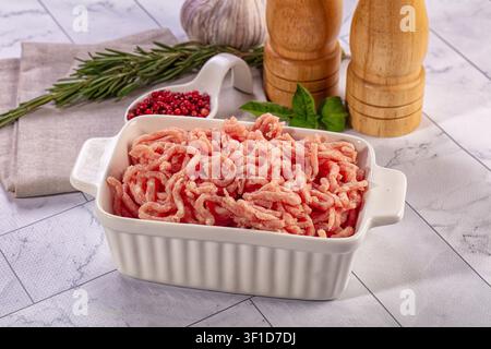 Fresh raw ground meat in white ceramic dish on marble surface. Composed with rosemary sprigs, garlic bulb, red peppercorns, basil leaves, wooden salt Stock Photo