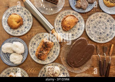 Assorted traditional pastries including empanadas, coconut balls, chocolate-covered cakes, and sprinkle-topped rolls arranged on blue and white floral Stock Photo