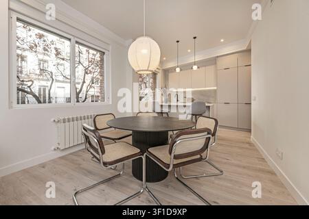 Contemporary open-plan dining and kitchen area featuring a round dark wood table with cane-back chrome chairs, large spherical pendant light, beige mi Stock Photo