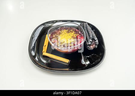 Traditional Peruvian purple corn mazamorra dessert served in a glass bowl with cinnamon sticks and dried purple corn cobs on a glossy black plate Stock Photo