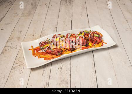 Grilled tandoori chicken pieces arranged on a sleek white rectangular plate with sautéed vegetables, topped with crisp onion rings, microgreens, and y Stock Photo