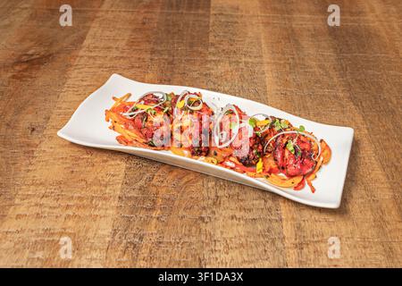 Spicy red marinated chicken pieces arranged on a long white rectangular plate with sautéed onions and peppers, garnished with microgreens, onion rings Stock Photo