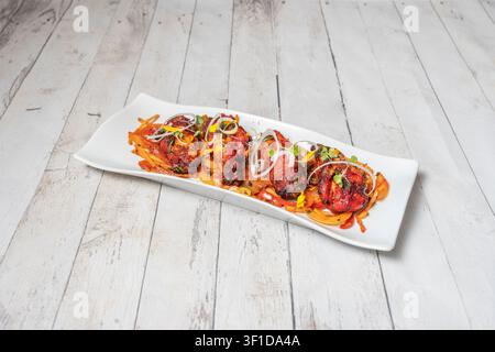 Spicy red marinated chicken pieces arranged on a long white rectangular plate with sautéed vegetables, topped with crisp onion rings, microgreens, and Stock Photo