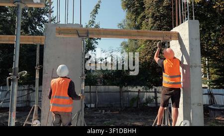 Construction Workers Installing Formwork on Concrete Walls. Two construction workers in safety gear adjust formwork between concrete walls at an Stock Photo