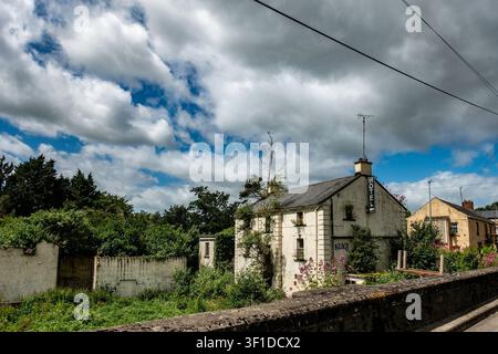 The derelict Old Mill Hotel building in Julianstown, Ireland, featuring overgrown vegetation, a hotel sign, and a stone wall under a cloudy sky. Stock Photo