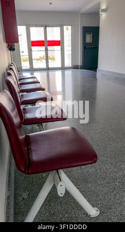 Brightly lit hallway showcases a series of empty red chairs lined up against a smooth floor. The sunlight filters through large doors, creating a calm Stock Photo