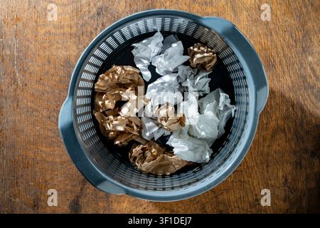Crumpled paper balls in office trash can, top view. White and brown crumpled paper balls in grey plastic waste basket on wooden desk representing brai Stock Photo