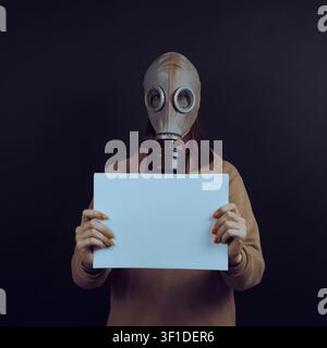 Woman in gas mask holding blank white paper banner. Girl wearing vintage gas mask holds empty white paper sheet on black background representing ecolo Stock Photo
