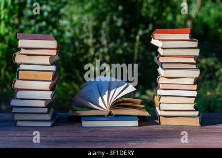 Two stacks of hardcover books flanking an open book with fanned pages on wooden table against blurred green nature background representing education a Stock Photo