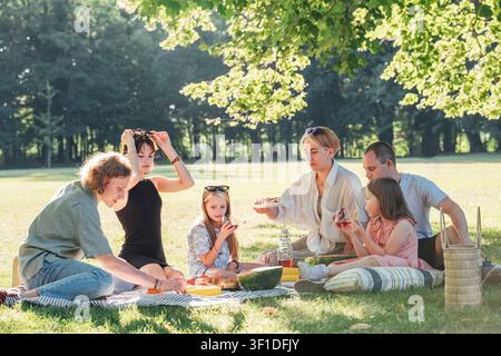 Large happy group of family and friends enjoying relaxing outdoor summer picnic under big green tree in sunny peaceful park eating fresh food and sitt Stock Photo