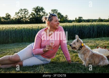 Carefree mature woman enjoying summer time with glass of wine and her dog. Outdoor relaxation and happiness Stock Photo