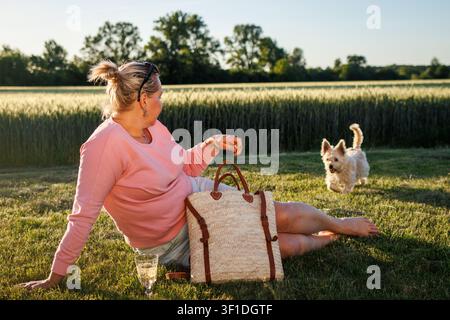 Plus size woman relaxing in summer rural scenery with glass of wine. Happy moment with dog outdoors Stock Photo