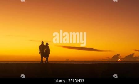 Silhouetted couple walking hand-in-hand on a coastal seawall at sunset, against a vibrant orange sky and sea horizon. Stock Photo