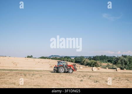 An agricultural tractor equipped with a bale spear standing in a harvested golden wheat field during summer season. Agriculture, France. Stock Photo