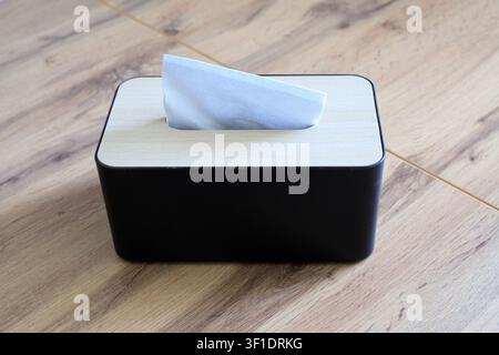 White paper napkin protrudes from a black tissue box. Napkin holder sits on a wooden table, top view. Modern hygiene concept in a home interior. Stock Photo