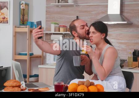 Happy young couple take selfies during breakfast in the kitchen, making funny faces and sharing a cheerful married lifestyle moment. Stock Photo