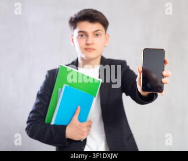 Guy in jacket with stack of notebooks shows empty mobile phone screen.Device close up unfocused Stock Photo