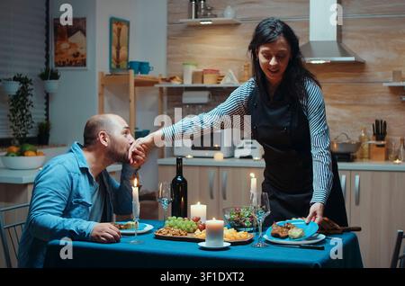 Thoughtful wife serves her husband during a romantic kitchen dinner, sharing healthy food and conversation at the anniversary table, inside a realistic lifestyle setting. Stock Photo
