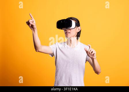 Asian man explores immersive virtual world, reaching out to interact with unseen digital elements. Modern male gamer enjoys futuristic entertainment while standing in studio with yellow background. Stock Photo