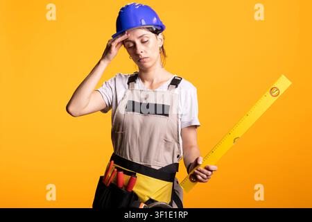 Female renovator wipes forehead in exhaustion while holding measuring level to represent fatigue or workload stress. Tired builder in safety helmet stands against yellow background wearing tool belt. Stock Photo