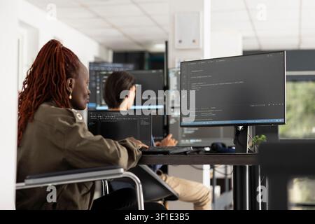 African American woman in wheelchair works at desktop PC, reviewing code and catching up on pending tasks. Injured female developer focuses on web development in professional office setting. Stock Photo