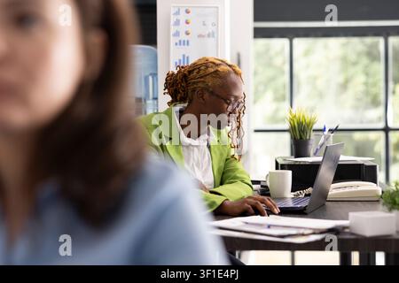 Businesswoman seated with focused posture and expression, dedicated in completing presentations and achieve company goals. Female manager analyzing information on laptop and organizing office tasks. Stock Photo