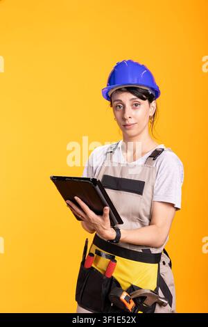 Skilled female tradesperson in hard hat using tablet to coordinate work activities. Woman renovator stands against yellow studio background, checking site data and digital project instructions. Stock Photo