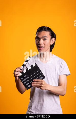 Excited man holds clapperboard toward camera over isolated background in studio. Male individual appears ready to start film scene, focused on work as director or producer during movie production. Stock Photo